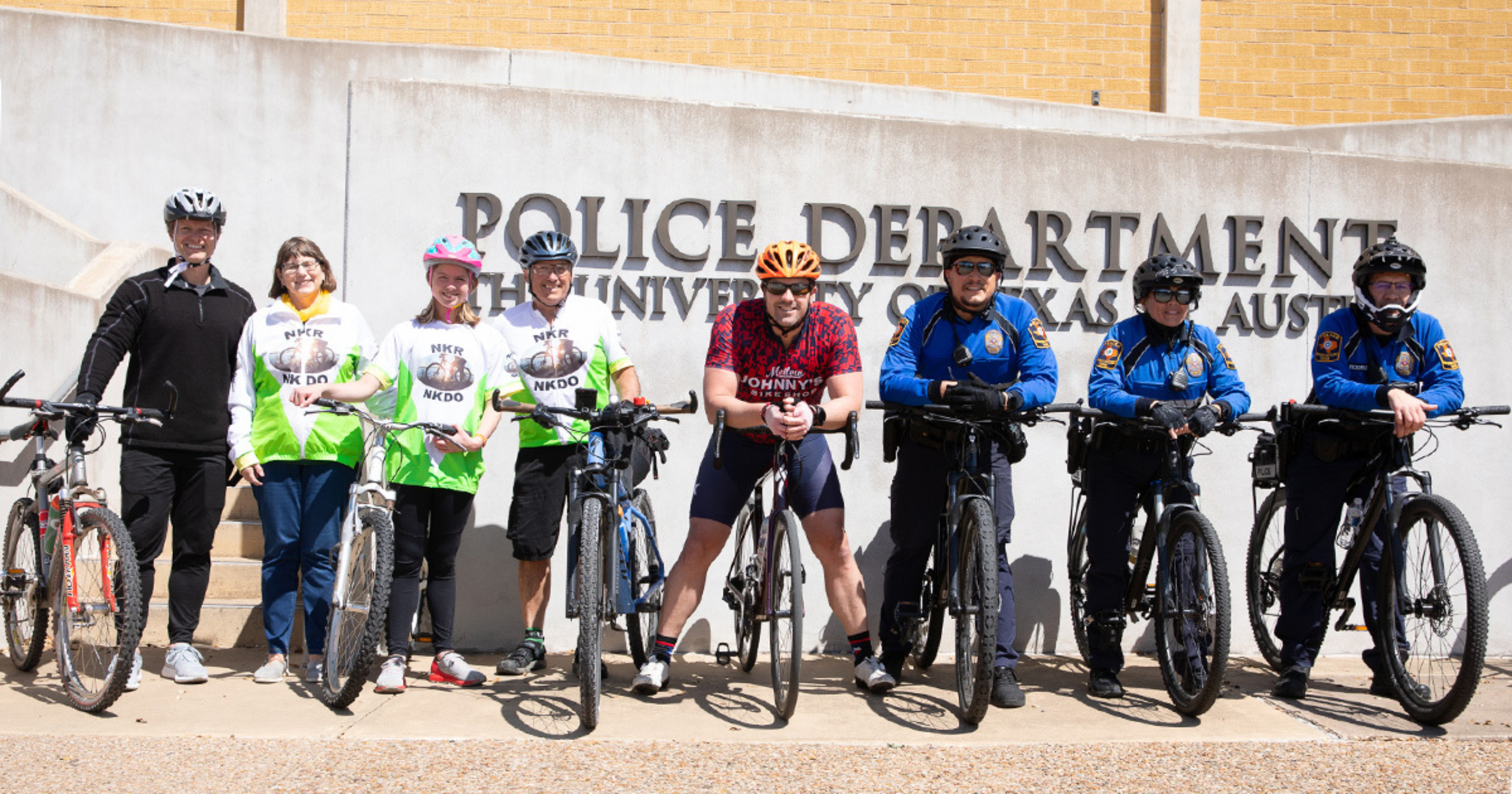 Travis Watson, Lynn Scotch, Rowan Sebek, Mark Scotch and Chance Sebek posing with members of the University of Texas Police Department outside of UTPD Headquarters. All those pictured except Lynn Scotch have bicycles beside them.