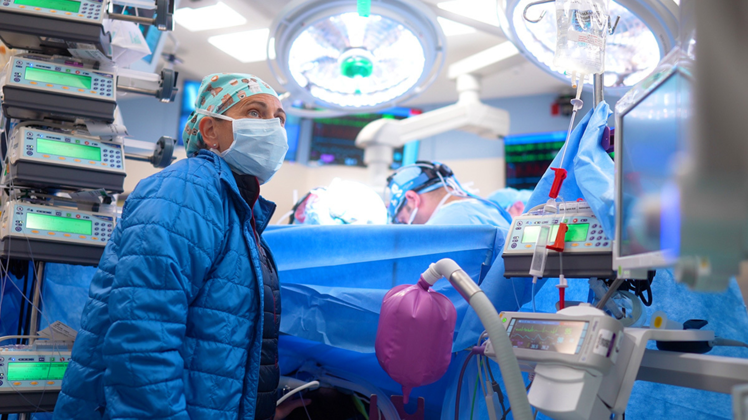 A female surgical nurse, masked with a blue coat monitors a patient, while a surgeon is working on the other side of the draped patient.