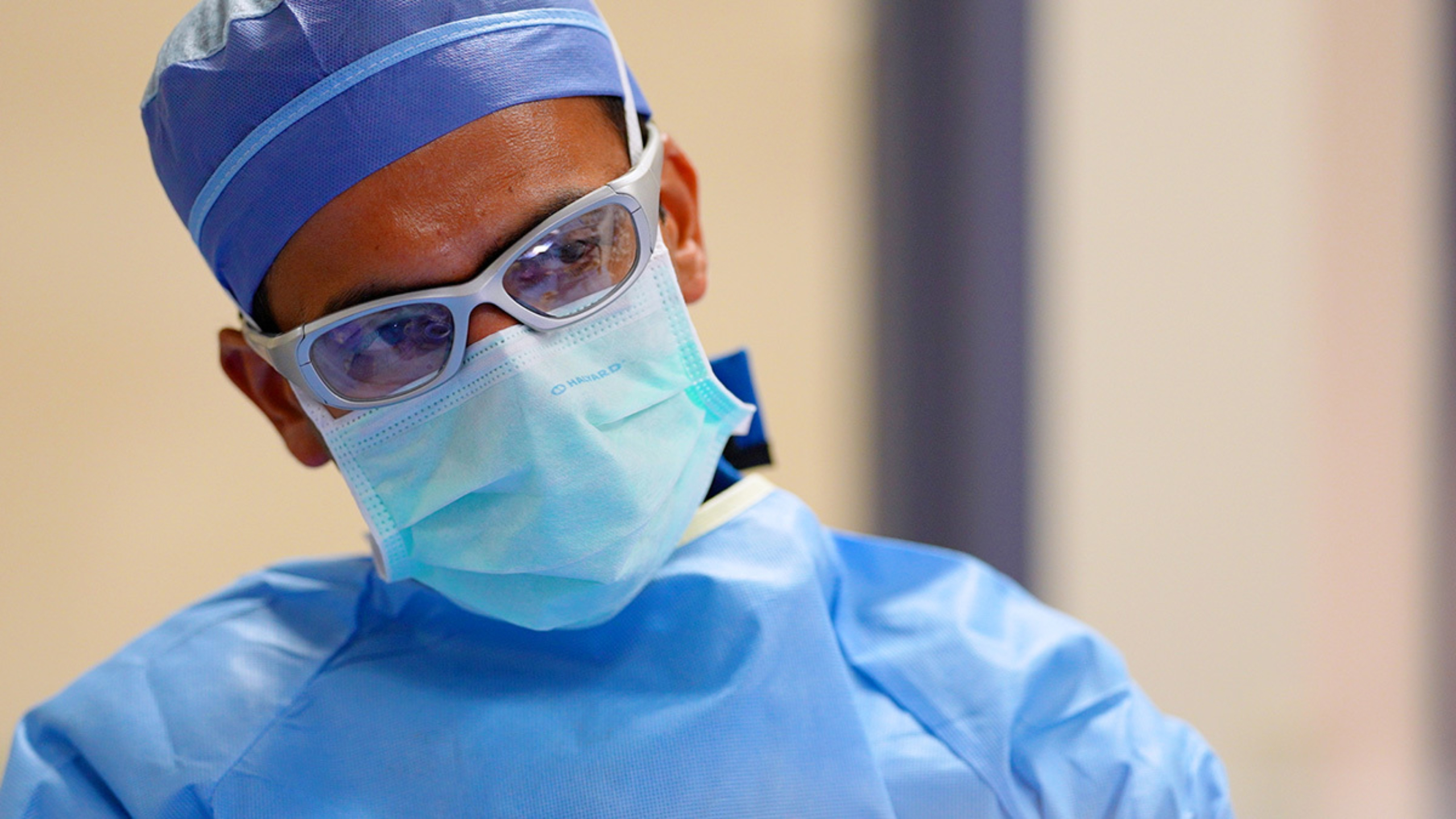 A male surgeon, masked with protective eyewear, tilts his head and looks at a monitor off screen.