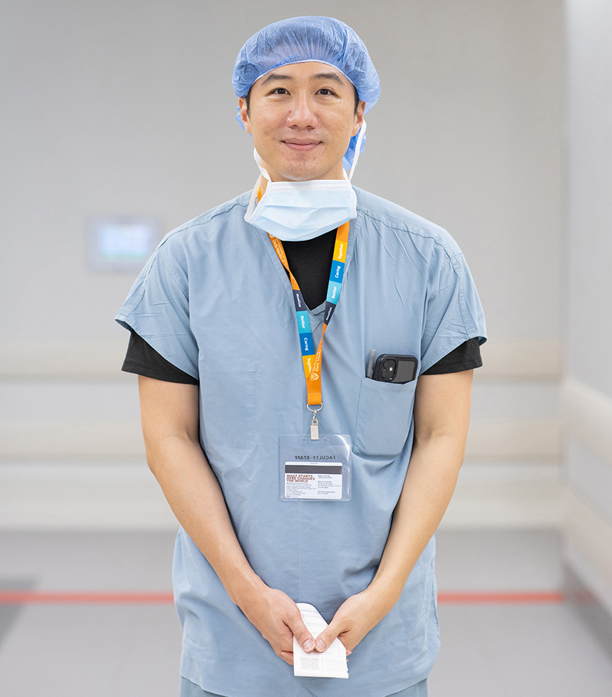 Ophthalmologist Gene Kim, MD stands in a surgical hallway dressed in scrubs and smiling.