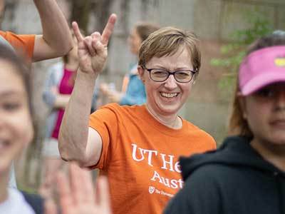 A woman standing outdoors in glasses and at UT Health Austin t-shirt smiles and makes a "Hook em" hand sign to the camera.