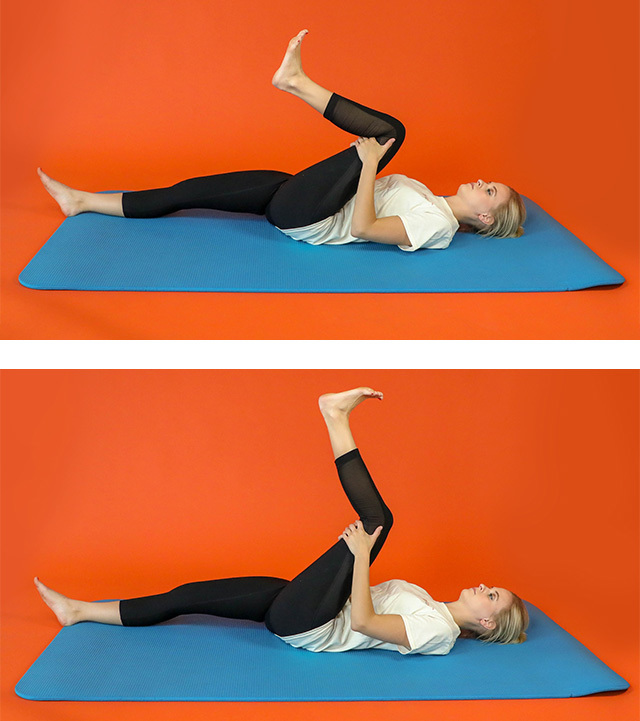 A woman lies on the floor, stretching the hamstring of her left leg by raising it in the air with her hands clasped behind her left knee.