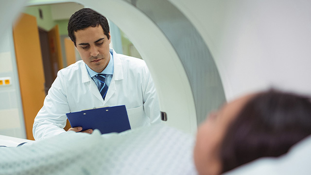 In this photo, taken from inside an MRI scan machine, a male clinician peers at a clipboard while a female patient waits inside the scanner.