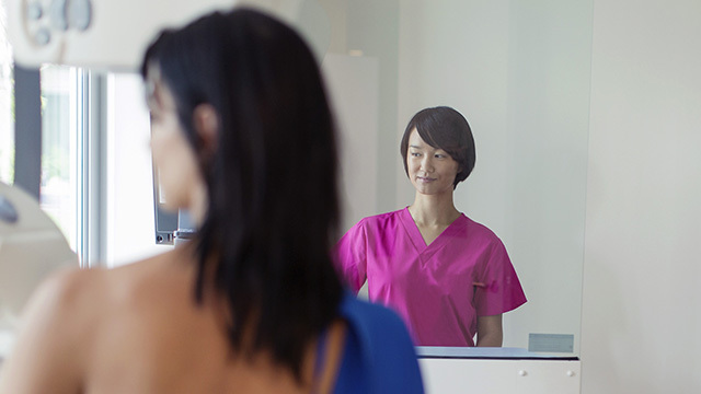 An Asian breast technician checks the screen while performing a mammogram on a patient.