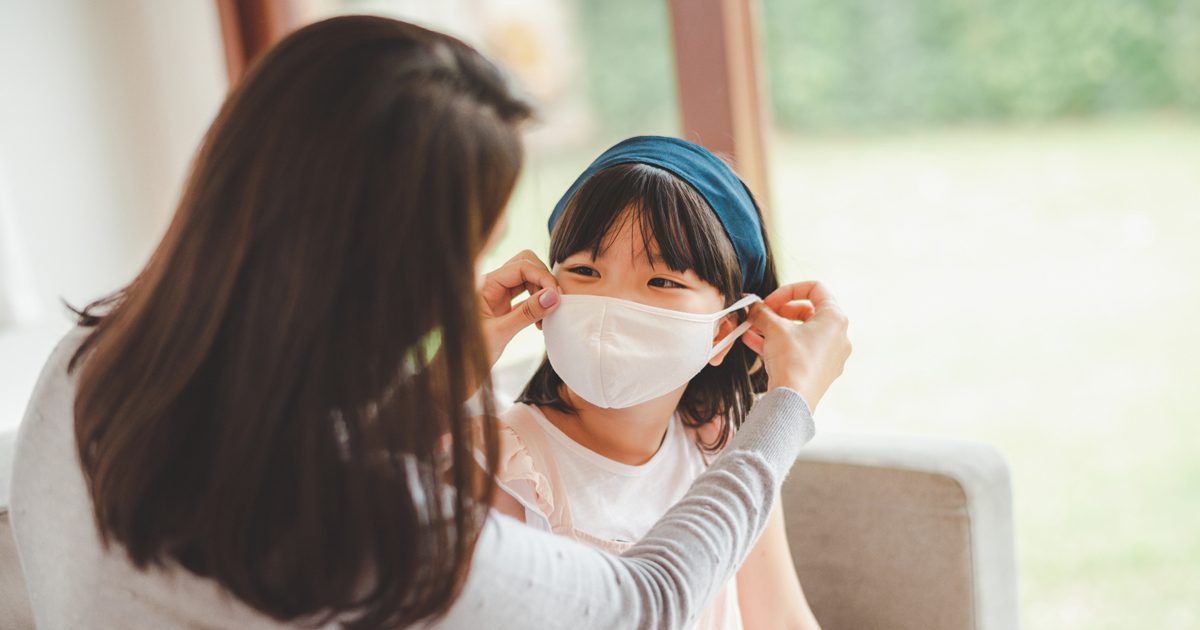 A mother putting a white cloth mask on a young girl wearing a blue headband.