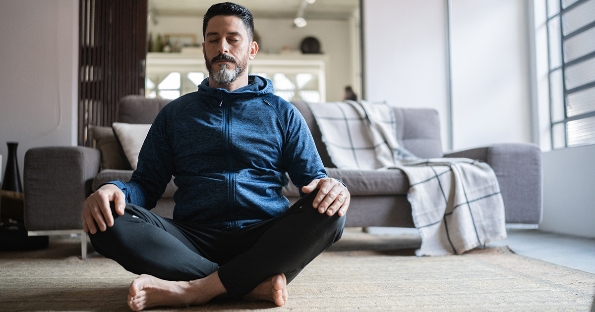 A white, middle aged man sits cross-legged in his living room practicing yoga or meditation.