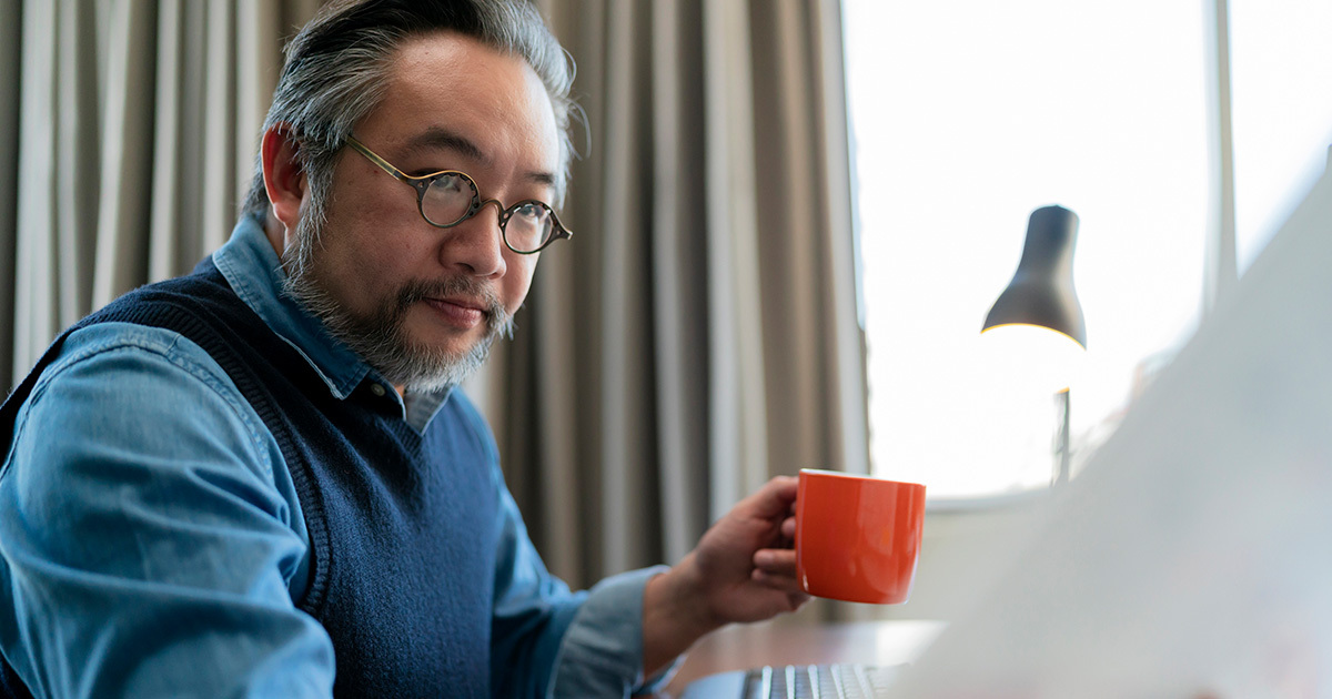 An East Asian, mature man with a beard and glasses, dressed in a blue button down shirt and nave blue sweater vest, glances to his left reading a document while holding an orange coffee mug in his right hand.