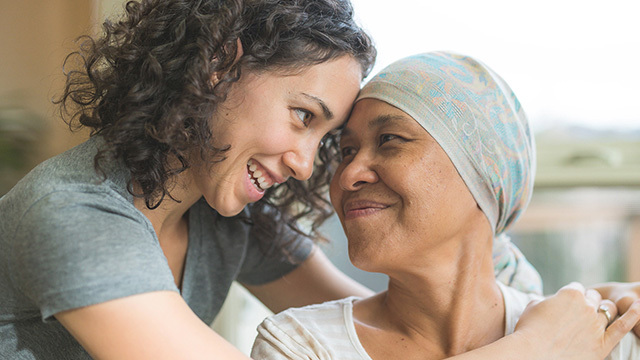 A young woman smiles at and embraces an older woman wearing a headscarf who is seated and looking at the younger woman.