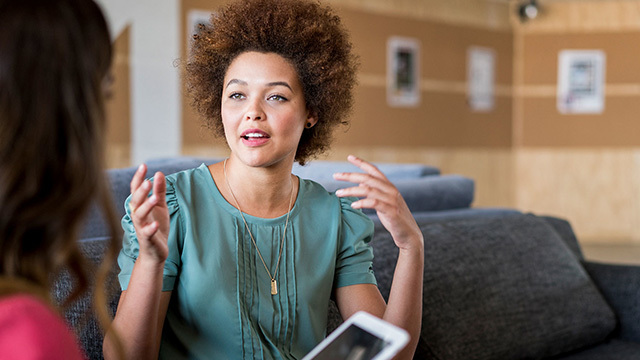 A young Black woman is facing and speaking to another woman to the left of the frame, while gesturing with her hands.