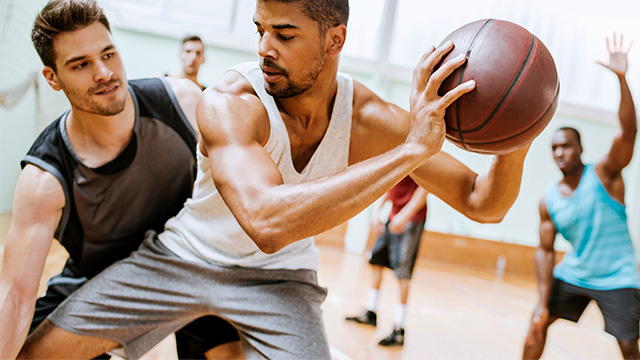 Two young adult males are playing basketball in a gym.