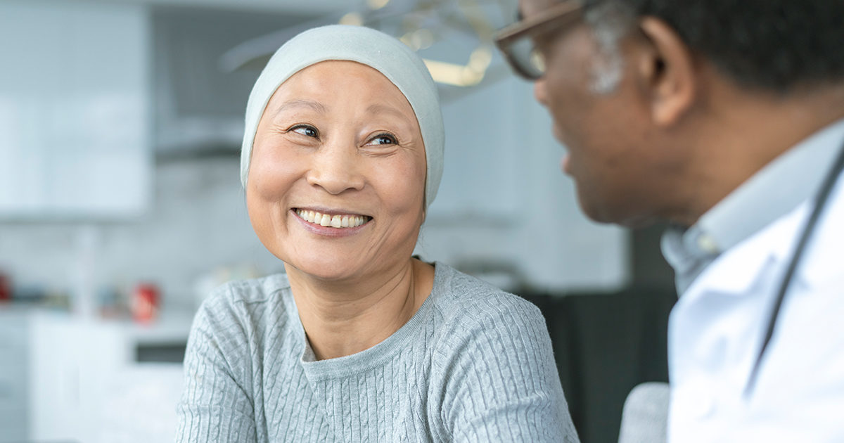 A mature Asian woman wearing a head scarf, smiles broadly while listening to a mature Black male health care provider.