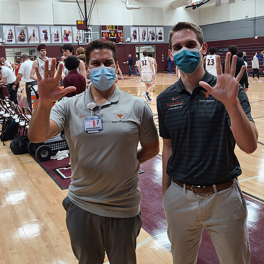 Two male doctors stand on the sidelines of a high school basketball court. Both are wearing masks to protect from COVID-19 infection. Both have their hands raised in greeting.