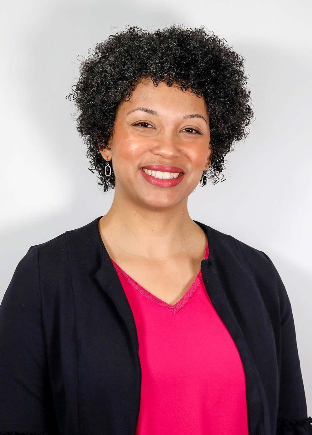 Donna Shanor wearing a pink blouse and black jacket and smiling in front of a white backdrop.