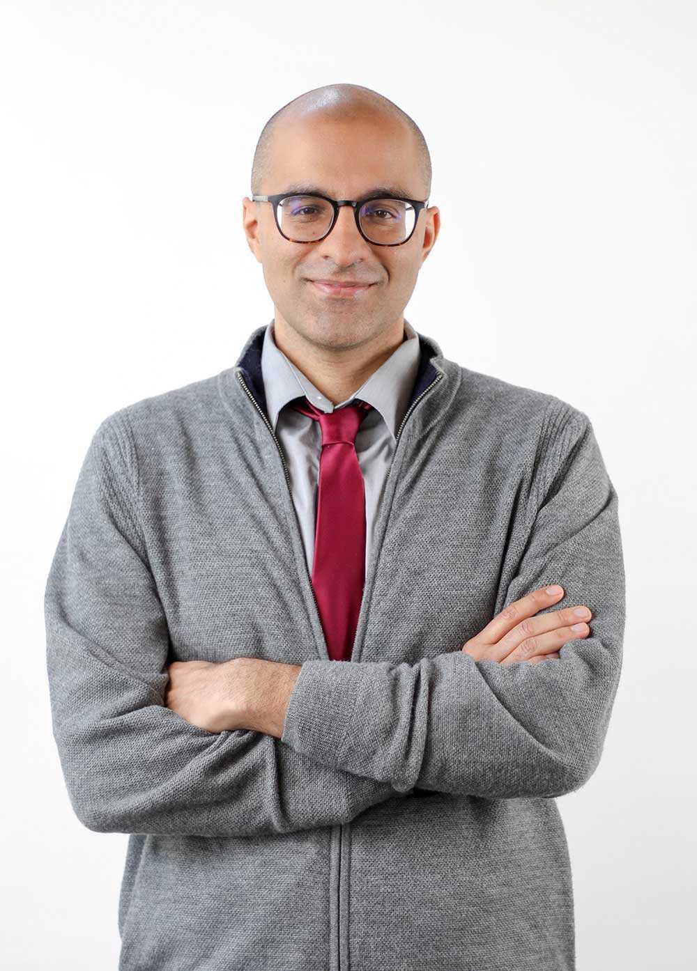 Jess Ambiee wearing a gray jacket and red tie and smiling in front of a white backdrop.