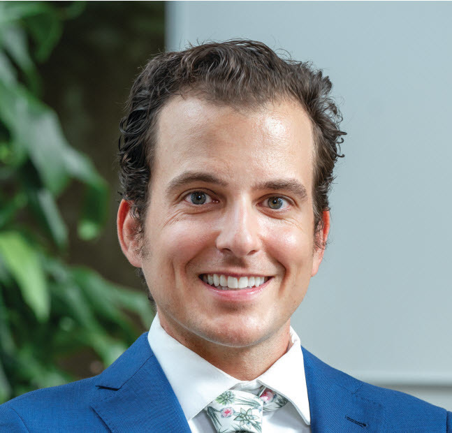 Urologic oncologist Aaron Lavinia, MD, MBA, wearing a suit and tie and smiling near a white wall and a tree.