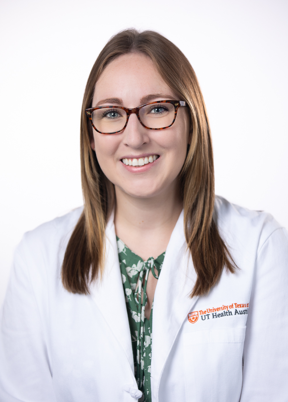 Genetic counselor Brynn Riley, MS, CGC, smiling in front of a white backdrop.  She is wearing glasses and a white coat with an embroidered UT Health Austin logo.