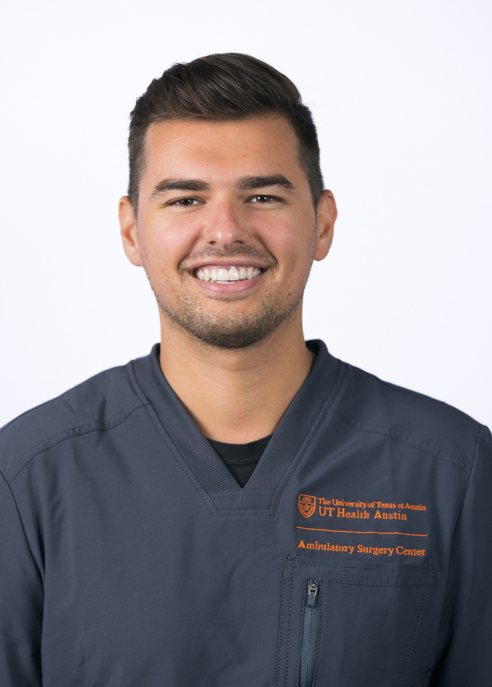 Christopher Ostrowski, Registered Nurse, is wearing charcoal gray scrubs and is smiling against a white background