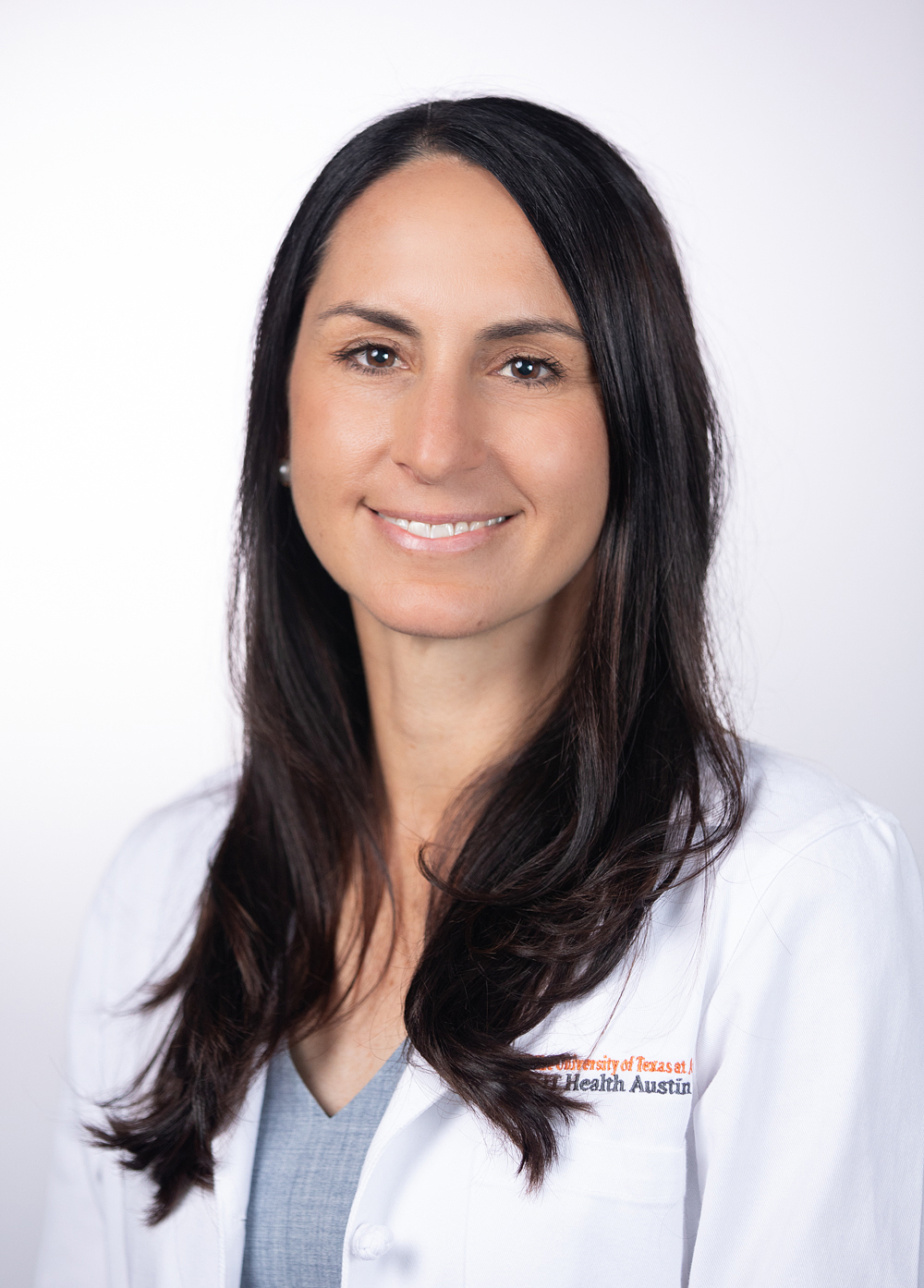 Nurse practitioner Elizabeth Whitten, MSN, APRN, FNP-C smiling in front of a white backdrop. She is wearing a white coat with an embroidered UT Health Austin logo.