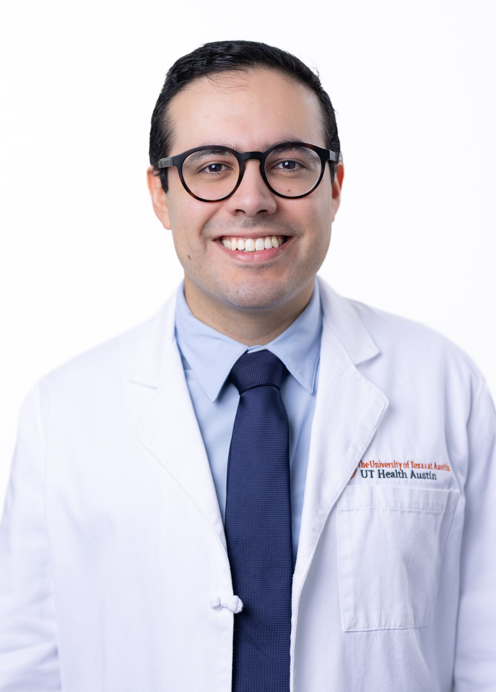 Felipe Flores is smiling at the camera. He is wearing a white coat, light blue shirt, and a navy blue tie.