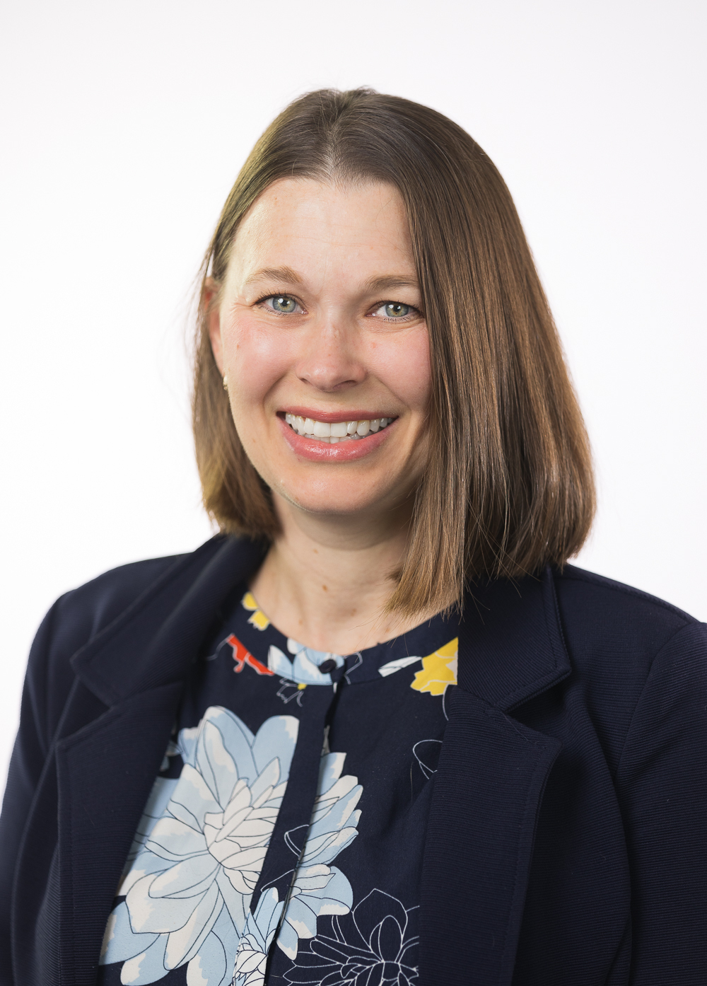 Social worker Jillian Bissar, smiling in front of a white backdrop. She is wearing a dark cardigan over a floral button down shirt,