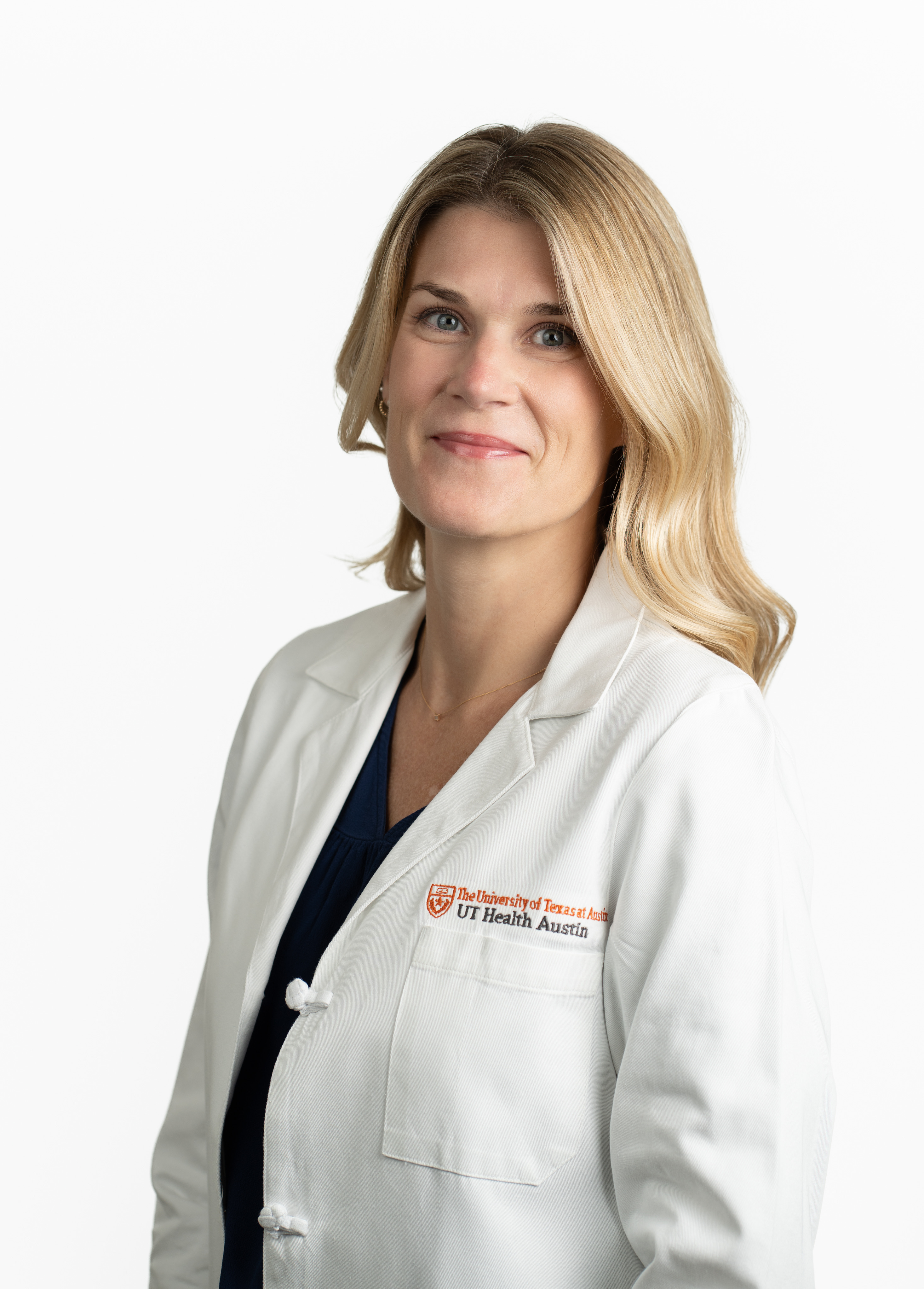 Nurse practitoner Kelly Swap, MSN, AGACNP-BC, AGNP-C, smiling in front of a white backdrop. She is wearing a white coat with an embroidered UT Health Austin logo.