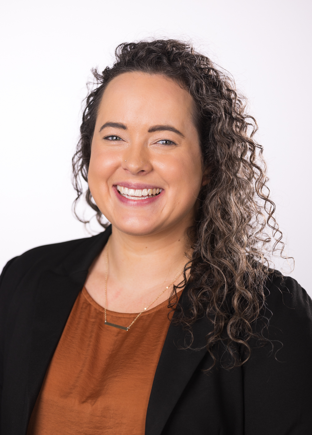 Rachel Holliman wearing an orange blouse and black blazer and smiling in front of a white backdrop.