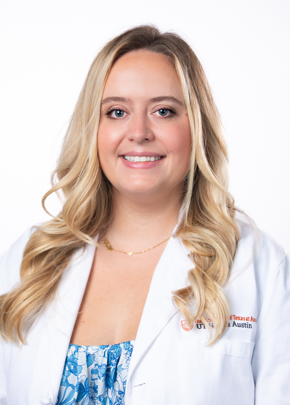 Nurse practitioner Stefanie Schutz MSN, FNP-C, CNOR, smiling in front of a white backdrop. She has long blond hair and is wearing white coat with an embroidered UT Health Austin logo.