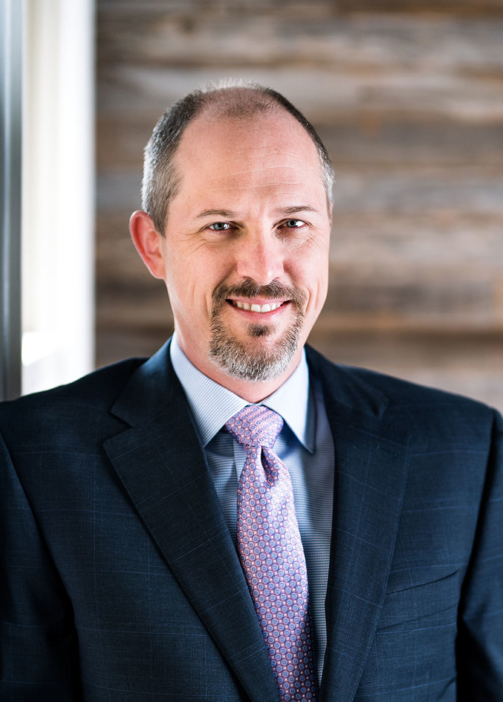 Dr. D. Byron Holt wearing a navy suit and patterned tie and smiling in front of a wooden wall.